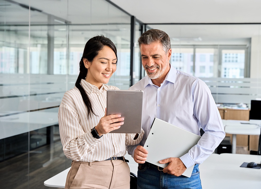 Employee Benefits - Portrait of a Smiling Young Asian Woman Showing a Middle Aged Male Coworker her Tablet as They Stand Inside a Modern Office
