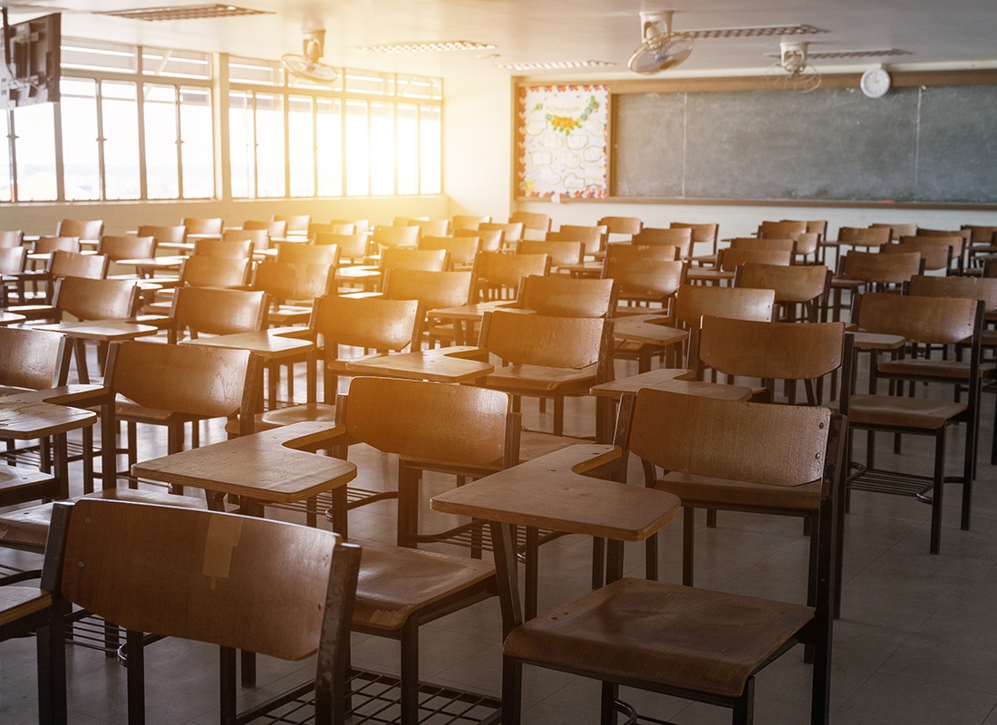 Insurance Solutions - View Inside a Classroom with Old Wooden Chairs and Desks with a Green Chalkboard in the Background and Sunlight Shining Through the Windows