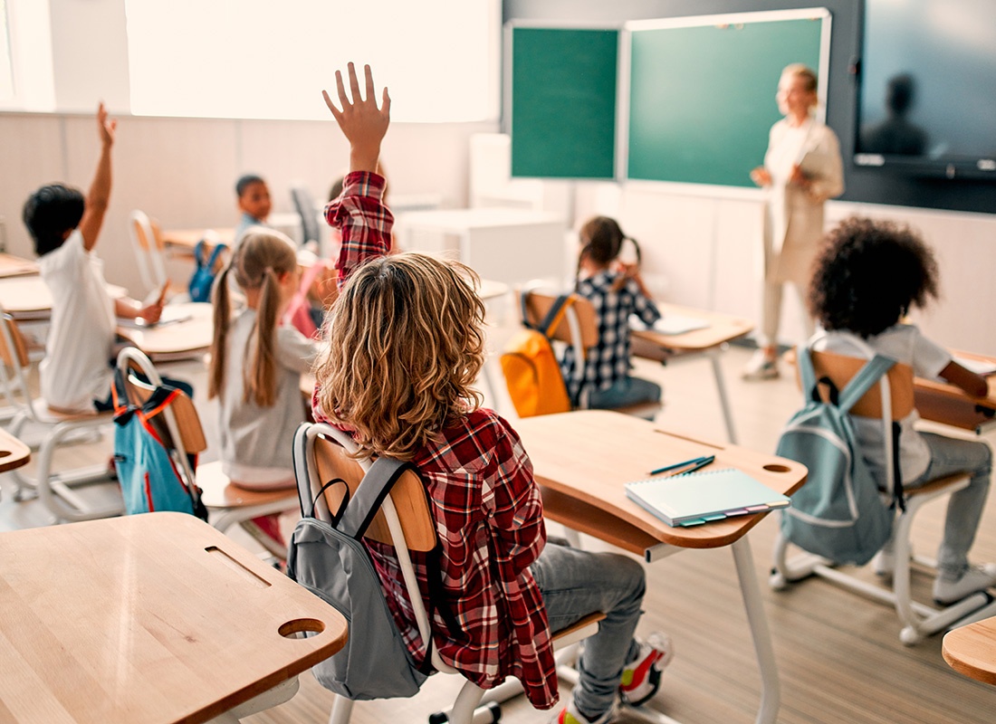 Insurance by Industry - View of a Group of Young Students Sitting in a Classroom Raising Their Hands with the Teacher Standing in Front Next to a Green Chalkboard